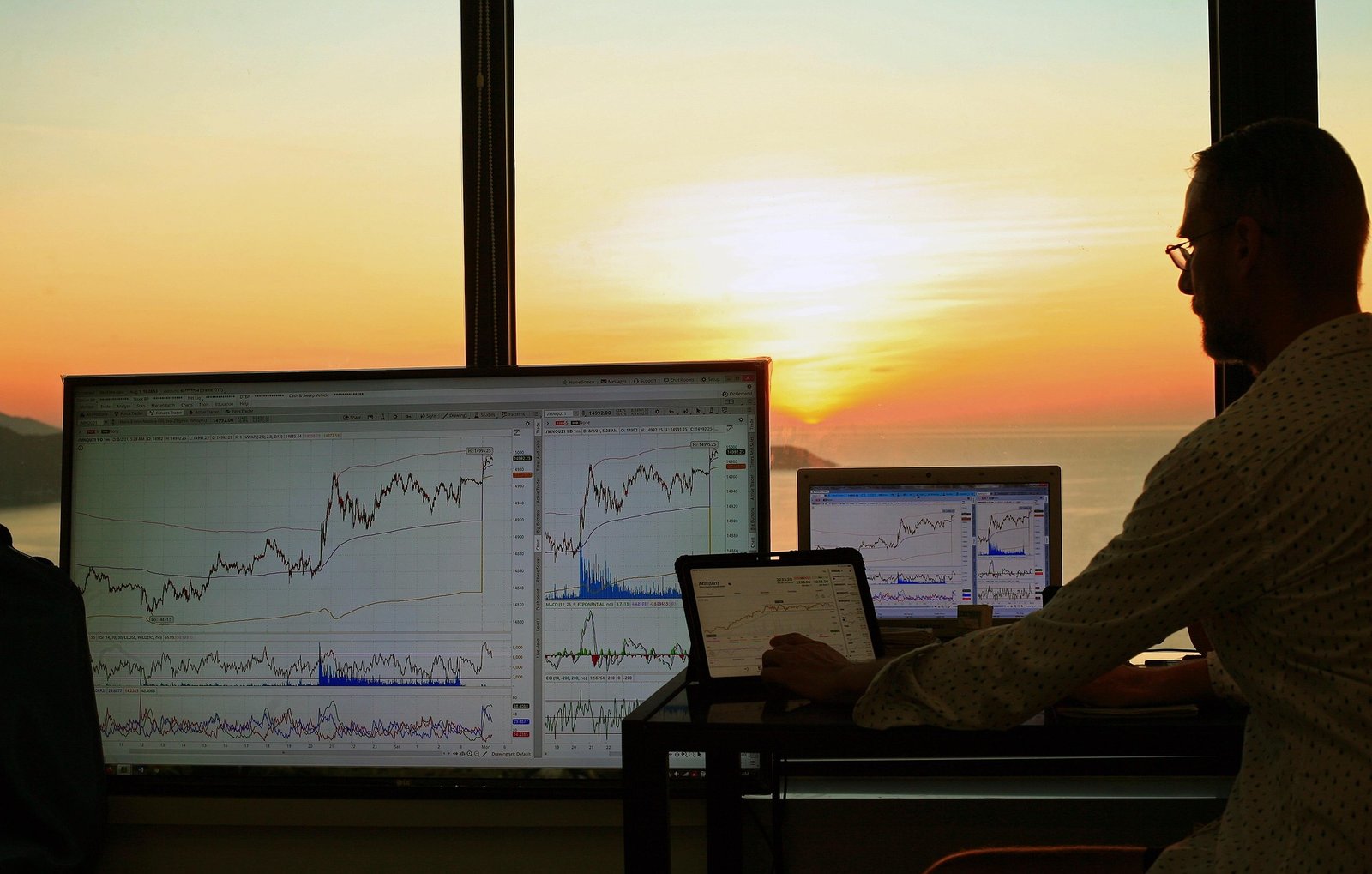 man analyzing forex trading charts on multiple screens during sunset with ocean view