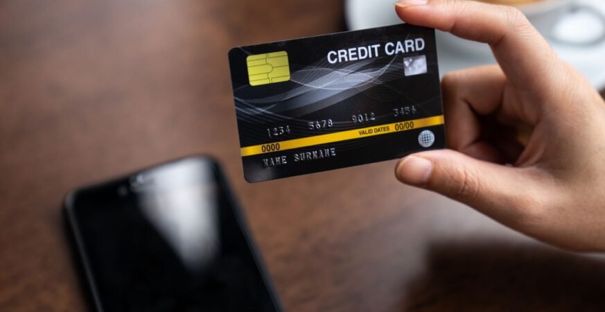 A person holding a black credit card above a wooden table with a smartphone and a coffee cup nearby, symbolizing online or card-based transactions.