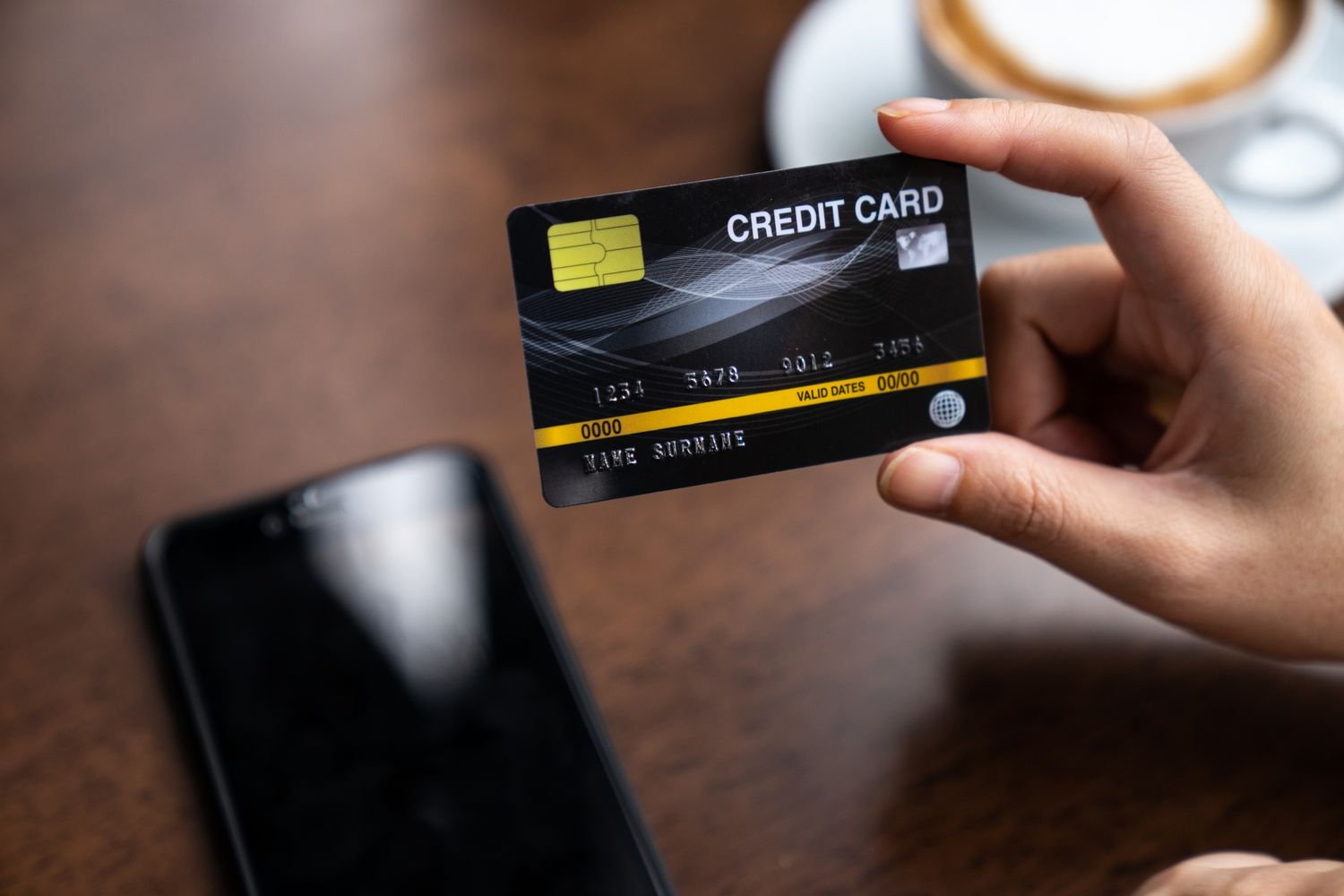 A person holding a black credit card above a wooden table with a smartphone and a coffee cup nearby, symbolizing online or card-based transactions.
